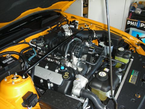 1998 Ford Crown Victoria - Open engine bay of a yellow car showing a black plastic intake manifold, accessory belts, and a yellow valve cover in a garage.