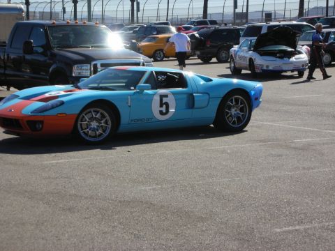 1998 Ford Crown Victoria - Heritage-livery Ford GT in light blue with orange stripes and a roundel number 5, parked in a racetrack lot among other cars.