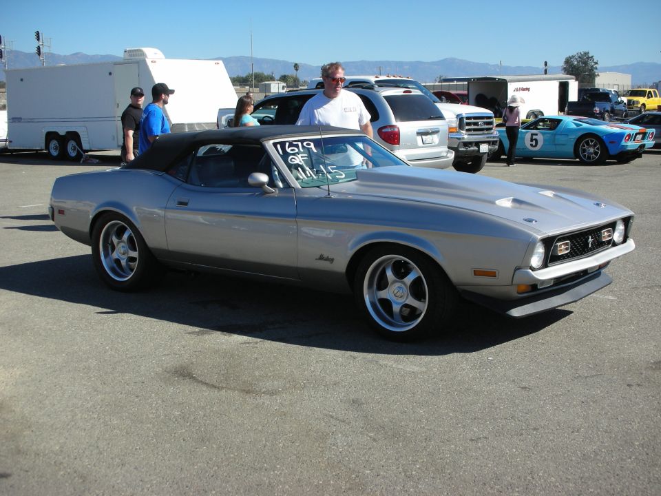 Silver Ford Mustang convertible with black top parked outdoors with people nearby