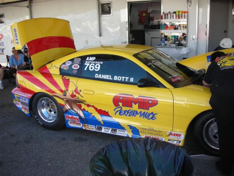 1998 Ford Crown Victoria - yellow drag-race Mustang lettered AMP Performance and Daniel Bott II, hood raised, sitting in a pit area.