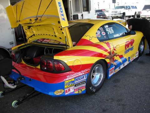 1998 Ford Crown Victoria - rear of a brightly painted drag-race Mustang on wheelie bars with its hatch open in a pit area.