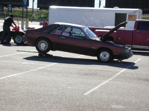 1998 Ford Crown Victoria - maroon Fox-body Mercury Capri hatchback drag car raised on stands with its hood open in a parking lot.