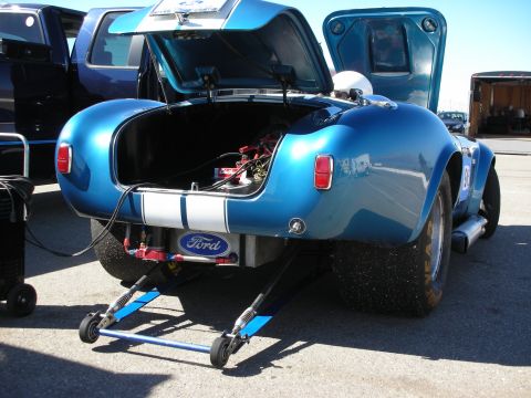 1998 Ford Crown Victoria - blue Cobra replica roadster with white stripes and a Ford-branded rear panel raised on jack stands outdoors.