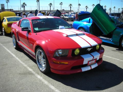 1998 Ford Crown Victoria - Red Ford Mustang GT with white racing stripes and chrome wheels parked among other cars at a sunny car show.