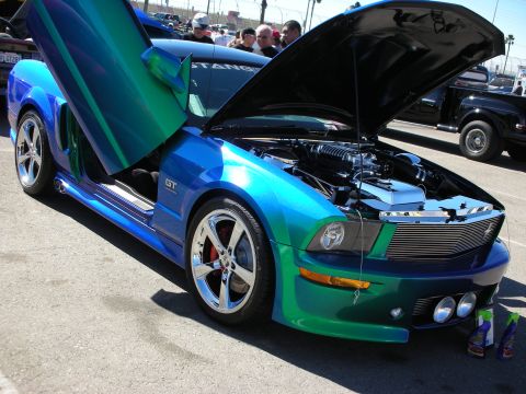 1998 Ford Crown Victoria - Color-shifting blue-green Ford Mustang GT with hood up and vertical doors open, on display at an outdoor car show.