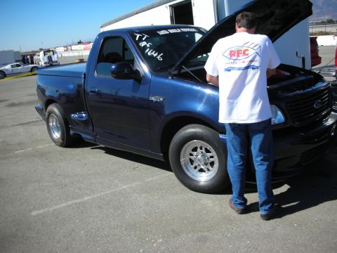 1998 Ford Crown Victoria - man in a white T-shirt leans into the open engine bay of a dark blue Ford regular-cab pickup parked beside trailers.
