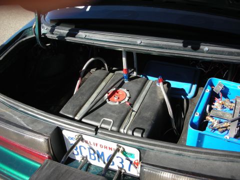 1998 Ford Crown Victoria - open car trunk fitted with relocated batteries, a black fuel cell, wiring, and a blue bin of parts above a California plate.