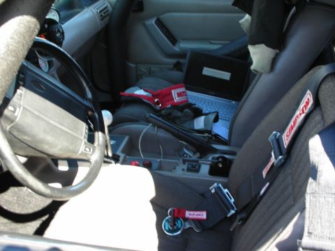 1998 Ford Crown Victoria - stripped race-car interior showing a worn steering wheel, a laptop on the seat, and Simpson racing harnesses.