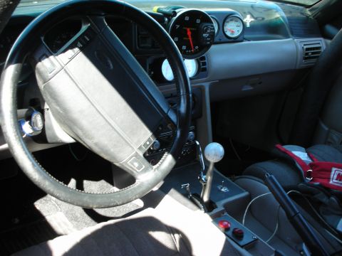 1998 Ford Crown Victoria - car interior with a Ford-badged steering wheel, a column-mounted tachometer, a B&M-style shifter, and a red racing harness.
