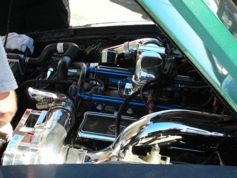 1998 Ford Crown Victoria - heavily chromed engine bay with twin polished intake pipes, blue fittings, and braided lines under the hood of a green car.