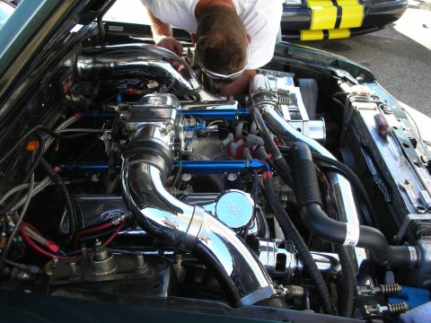 1998 Ford Crown Victoria - man leans into a chromed engine bay full of polished pipes and blue fittings, with a yellow-striped car visible behind him.