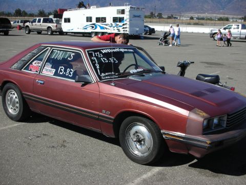 1998 Ford Crown Victoria - maroon 1980s Mercury Capri 5.0 fastback with race numbers on the glass parked at a desert drag strip near an RV.