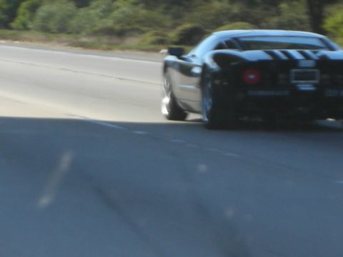 1998 Ford Crown Victoria - black two-seat sports car with racing stripes accelerates along an open road in a motion-blurred rear three-quarter shot.