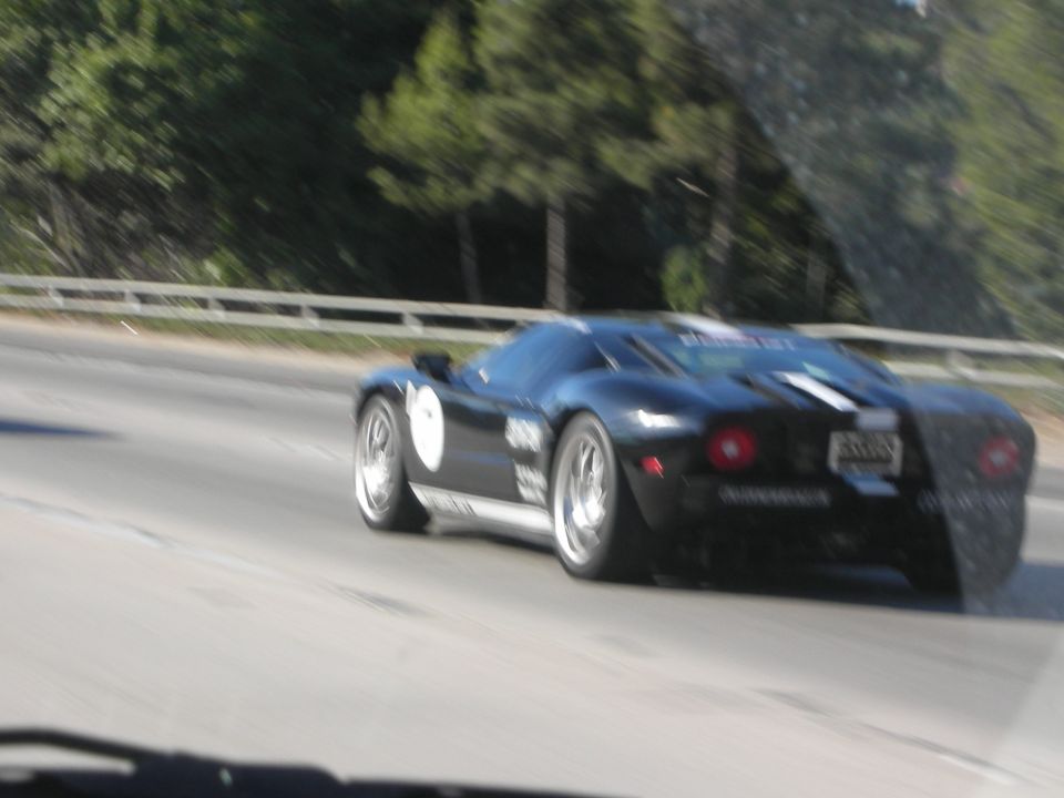 Black sports car with white stripes and circular taillights driving past a guardrail