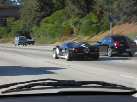 1998 Ford Crown Victoria - black striped sports car overtakes a black sedan on a multi-lane highway, seen through a windshield with wiper visible.