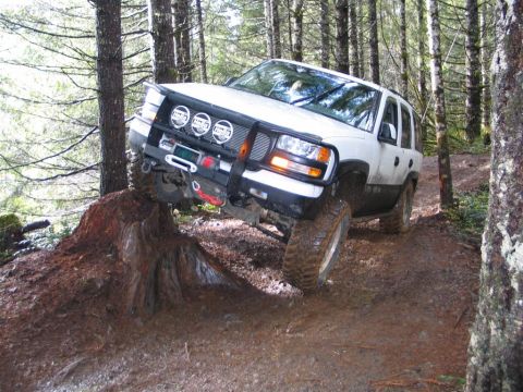 2002 GMC Yukon - White SUV navigates a challenging off-road course, climbing over a large tree stump.