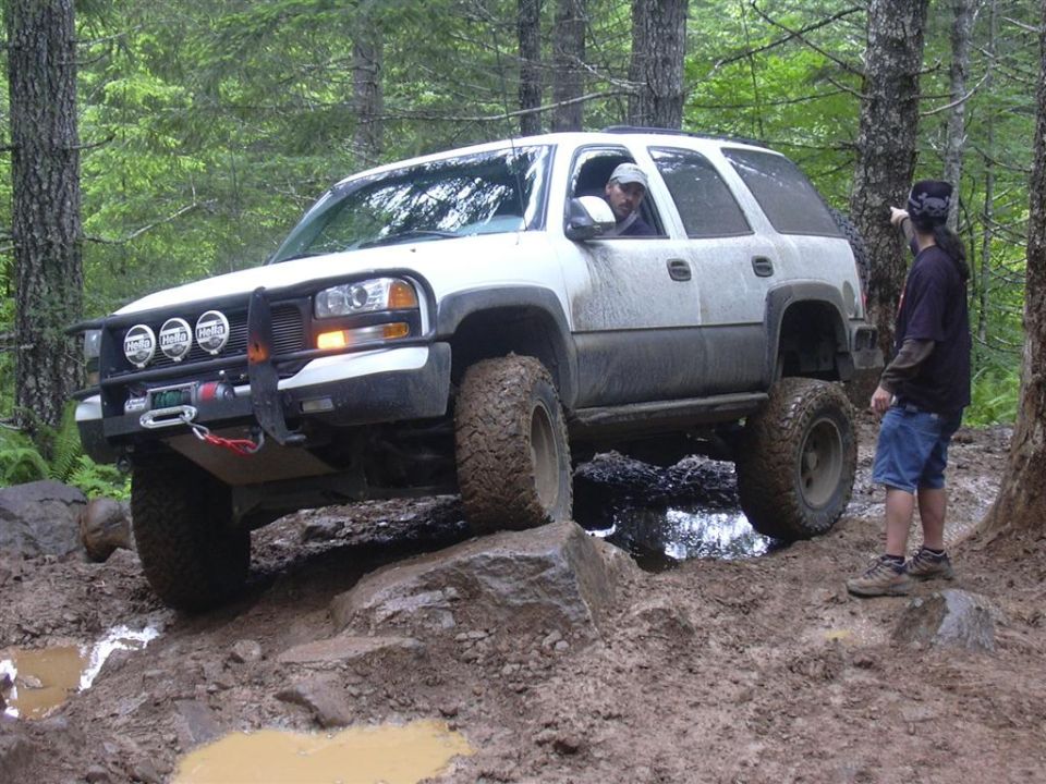 Lifted white full-size SUV with brush guard, Hella lights, and winch crawling a muddy forest trail