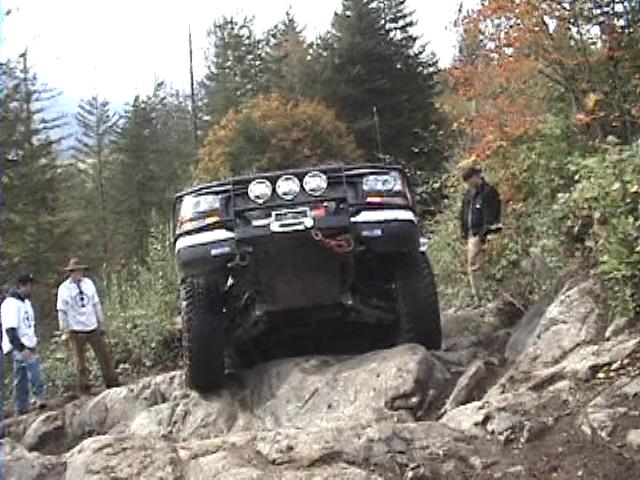 Lifted dark SUV with winch and light bar climbing a rock ledge on a forest trail
