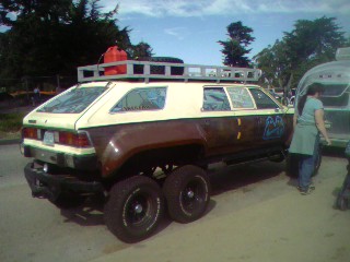 1994 Ford Bronco - Rear three-quarter view of a heavily modified six-wheeled art car built from a wagon body, painted brown and cream with graffiti-style lett…