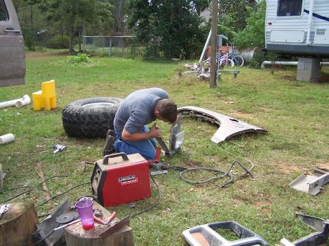 1981 Ford Bronco - Man works on a car fender and headlight with a welder, outdoors.