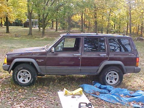 1991 Ford Bronco - A maroon Jeep Cherokee is parked outdoors on a grassy area with trees in the background.