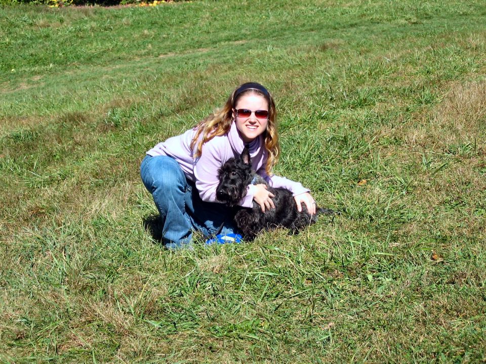 Woman crouching on grass, hugging a black Scottish Terrier