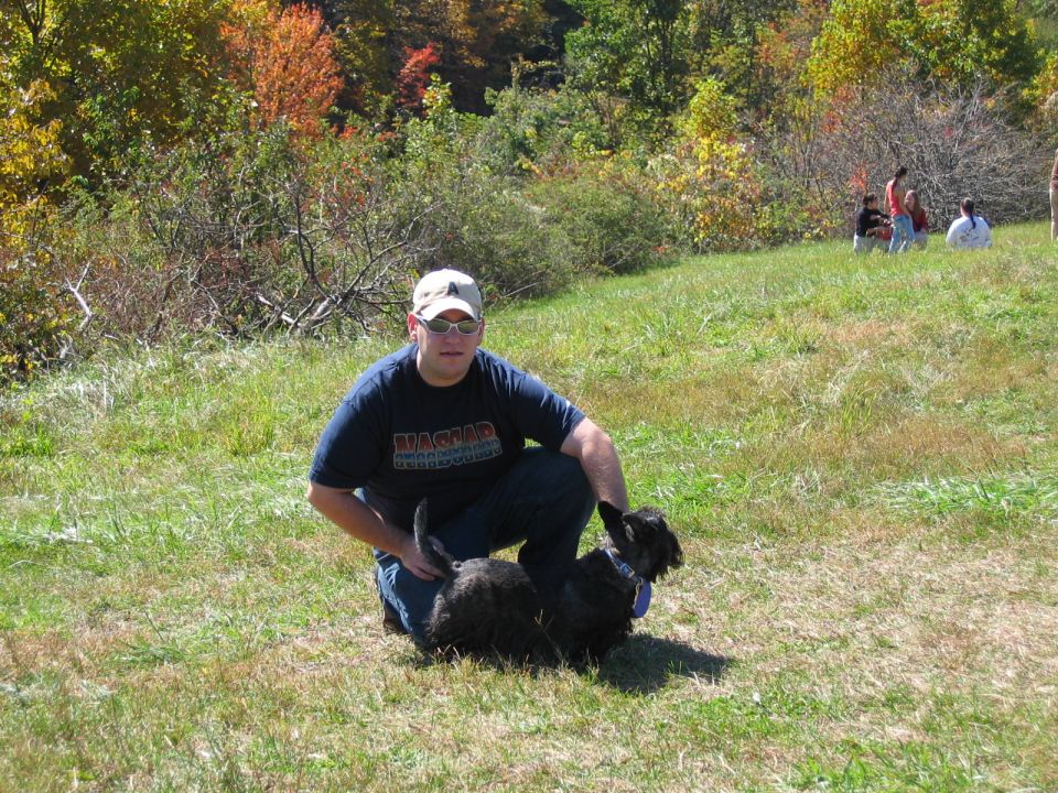 Man wearing a dark blue t-shirt and a baseball cap kneels next to a black Scottish Terrier in a grassy field. Trees with fall colors are in the background