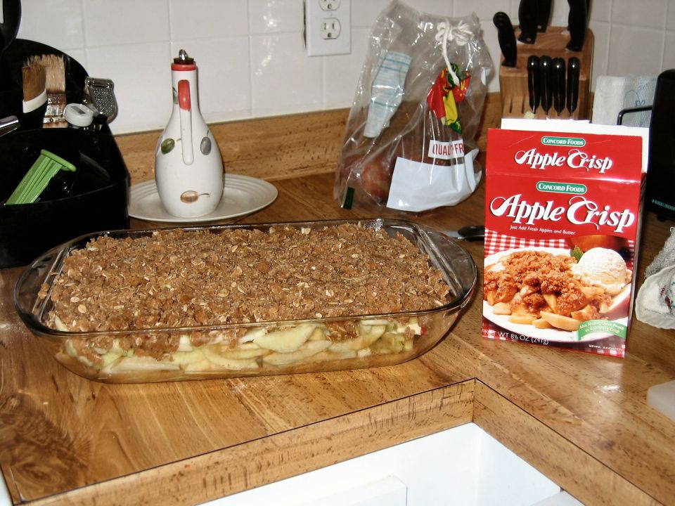 Unbaked apple crisp in a glass dish next to a box of Concord Foods Apple Crisp mix on a wooden countertop
