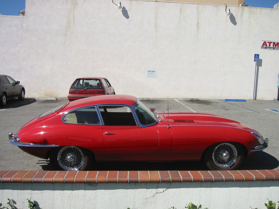 Red Jaguar E-Type coupe side profile on chrome wire wheels in a parking lot