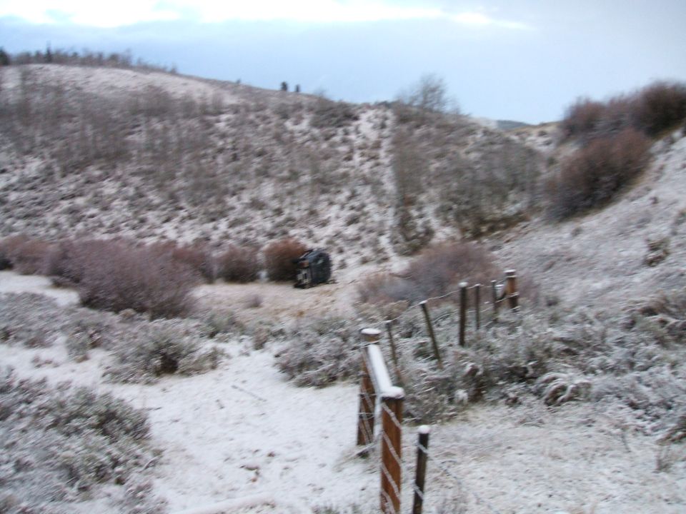 Overturned dark-colored vehicle in a snowy, brush-covered landscape with a fence in the foreground