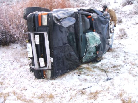 1992 Ford Ranger - A black truck lies on its side in a snowy, rural area, with a person standing nearby.
