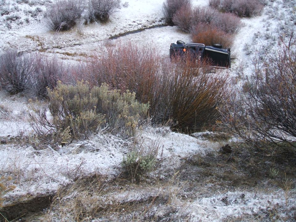 Overturned black truck lying on its side in a snowy ditch with brush surrounding it
