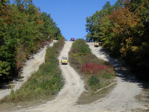 2005 Jeep Wrangler - Multiple off-road vehicles ascend a steep, wooded dirt track under a clear blue sky.