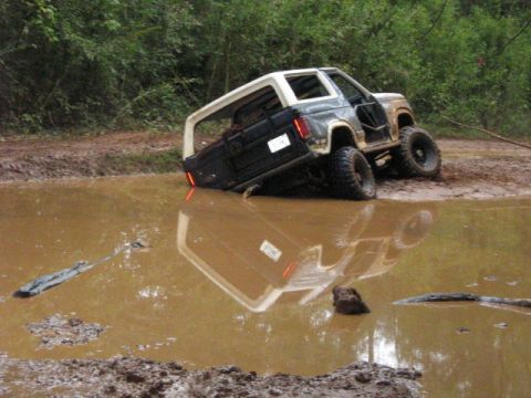 1986 Ford Bronco - A Ford Bronco is stuck in a deep mud puddle with its rear end submerged and the driver's door open.