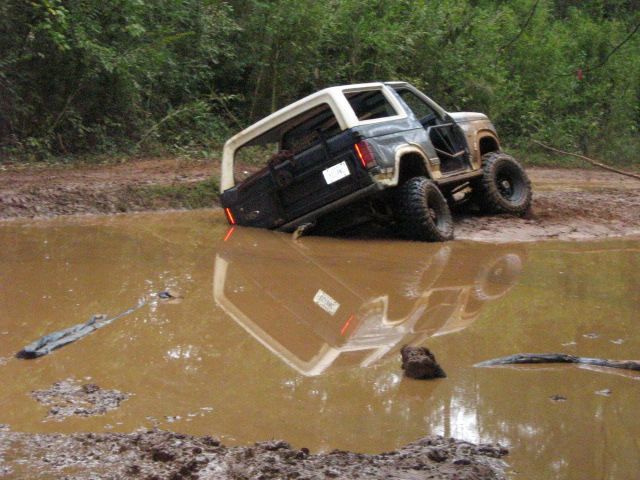 Dark blue and white Ford Bronco stuck in a mud puddle, driver's door open, rear end submerged
