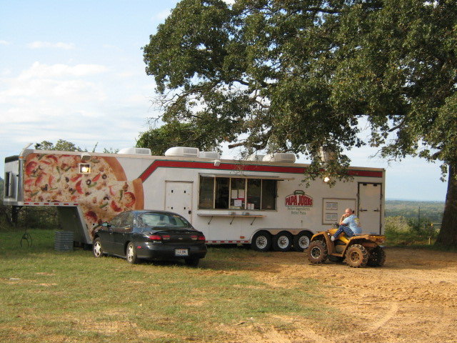 Papa John's pizza trailer with a sedan and ATV parked nearby under a large tree