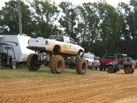 1986 Ford Bronco - Modified Dodge pickup truck with oversized tires at an outdoor event, alongside a trailer and other vehicles.