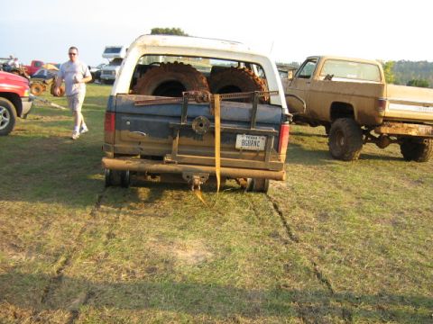 1986 Ford Bronco - Muddy Ford Bronco loaded with spare tires, parked next to a tan pickup truck in a grassy field.