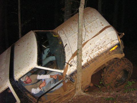 1986 Ford Bronco - A white off-road vehicle is stuck in trees and covered in mud, with a person inside pointing.