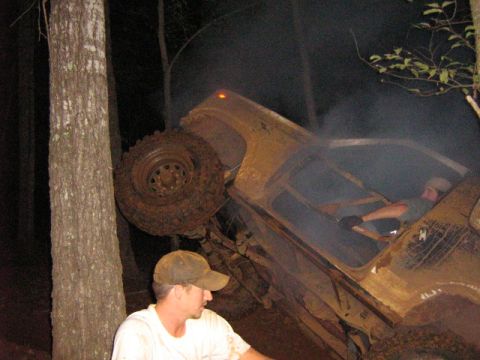 1986 Ford Bronco - An overturned SUV, likely from the 1980s, lies on its side in a wooded area, emitting smoke.