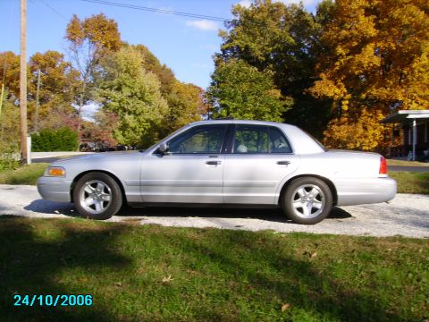2002 Ford Crown Victoria - A silver 2002 Ford Crown Victoria sedan is parked outdoors on a sunny day with autumn foliage in the background.