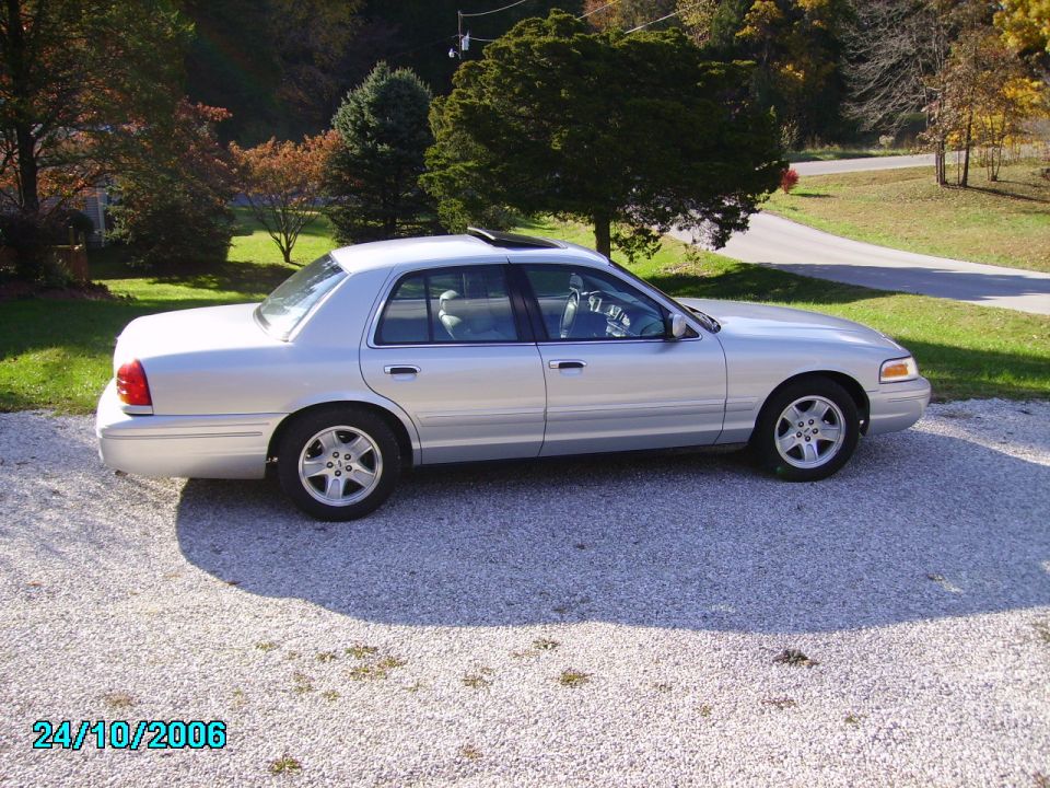 Silver 2002 Ford Crown Victoria parked on gravel with trees and a road in the background