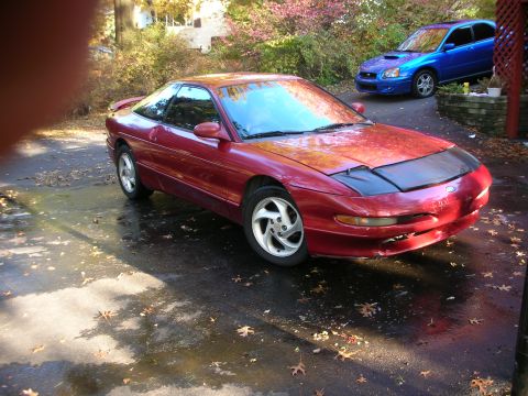 1995 Ford Probe - A red 1995 Ford Probe is parked on a wet driveway, with a blue Subaru in the background.