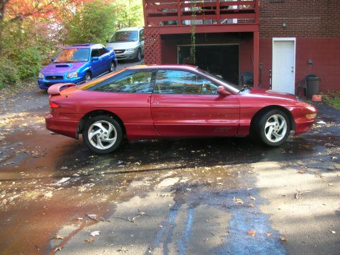 1995 Ford Probe - A red 1995 Ford Probe is parked in a driveway next to a blue Subaru and a silver van.