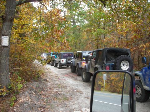 1997 Jeep TJ - Multiple 1997 Jeep TJs drive in a line on a dirt road surrounded by autumn foliage.