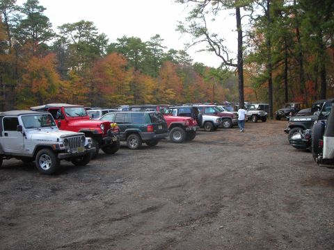 1997 Jeep TJ - A collection of Jeep Wranglers and SUVs are parked in a dirt lot surrounded by trees displaying fall colors.