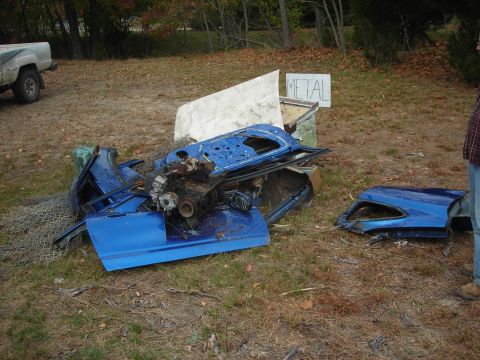 1997 Jeep TJ - Scattered blue car body panels and engine components are piled up in a yard with a 'METAL' sign in the background.