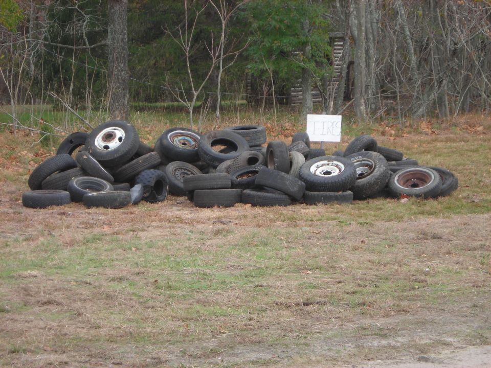 Pile of used tires on grassy ground with trees in background. A white sign with text is visible