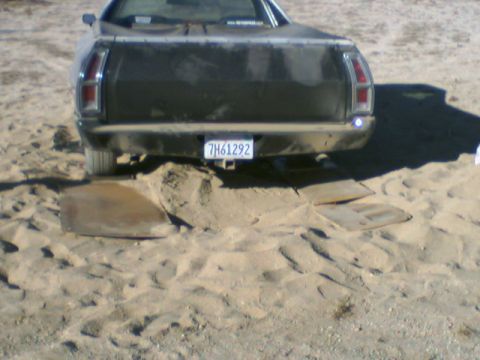 1972 Ford Ranchero - A dark 1972 Ford Ranchero is partially buried in sand, with wooden planks under its rear wheels.