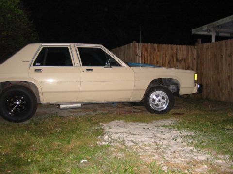 1985 Ford LTD Crown Victoria - Side view of a tan sedan with custom wheels and exhaust pipes, parked in a yard at night.